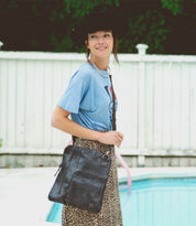 A woman stands by a pool, smiling and carrying the Bed|Stü Aiken full-grain leather bag with an adjustable strap, paired with a blue T-shirt, leopard print skirt, and black hat.