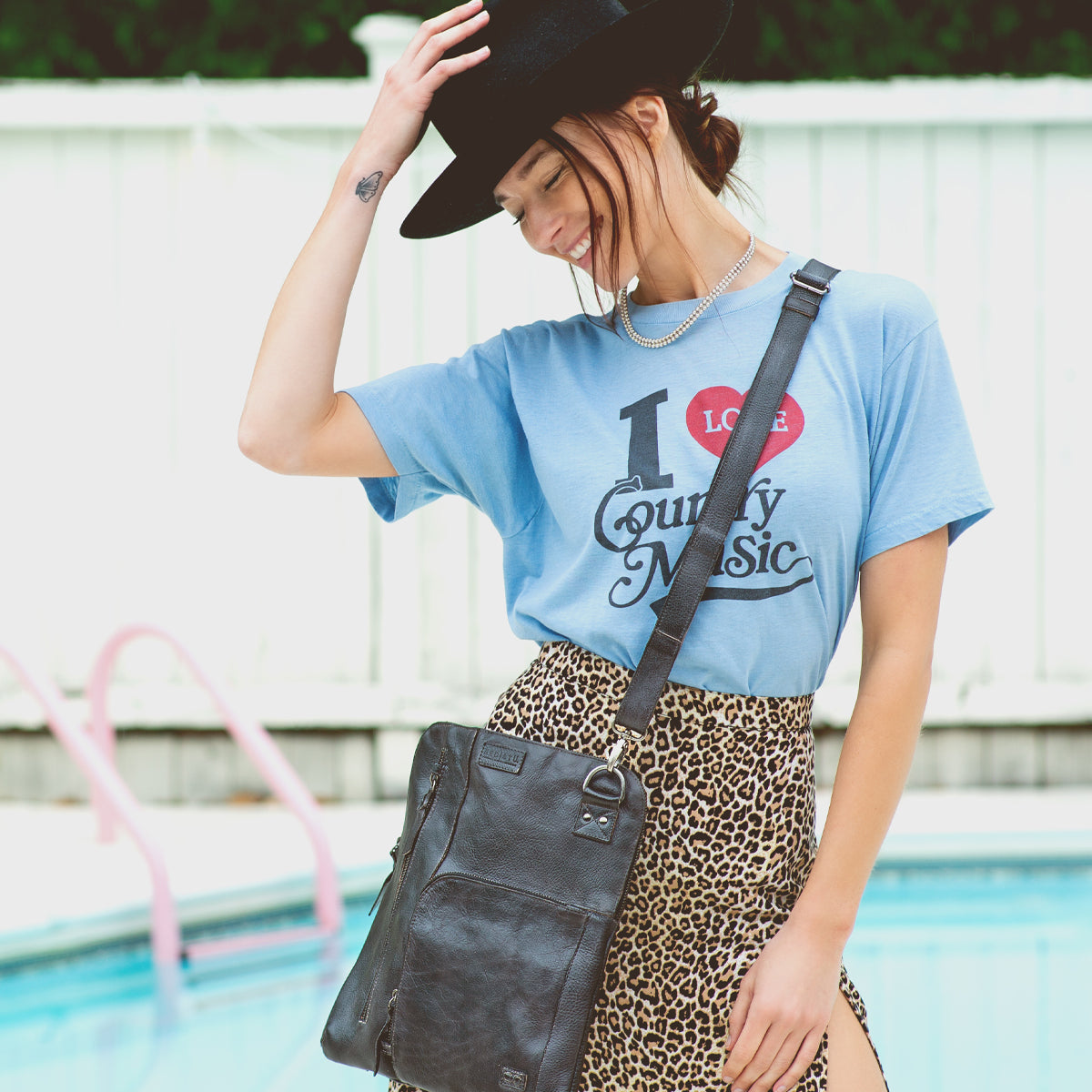 By the pool, a woman smiles with her hand on her hat, wearing a black hat, blue "I Love Country Music" shirt, leopard print skirt, and the Bed|Stü Aiken crossbody bag crafted from full-grain leather with an adjustable strap.