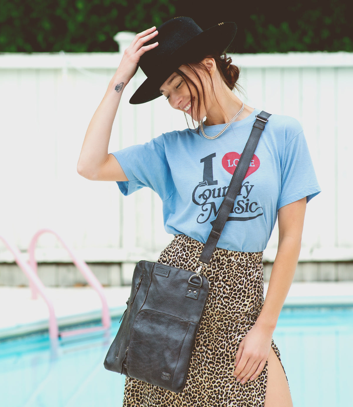By the pool, a woman smiles with her hand on her hat, wearing a black hat, blue "I Love Country Music" shirt, leopard print skirt, and the Bed|Stü Aiken crossbody bag crafted from full-grain leather with an adjustable strap.