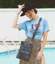 By the pool, a woman smiles with her hand on her hat, wearing a black hat, blue "I Love Country Music" shirt, leopard print skirt, and the Bed|Stü Aiken crossbody bag crafted from full-grain leather with an adjustable strap.