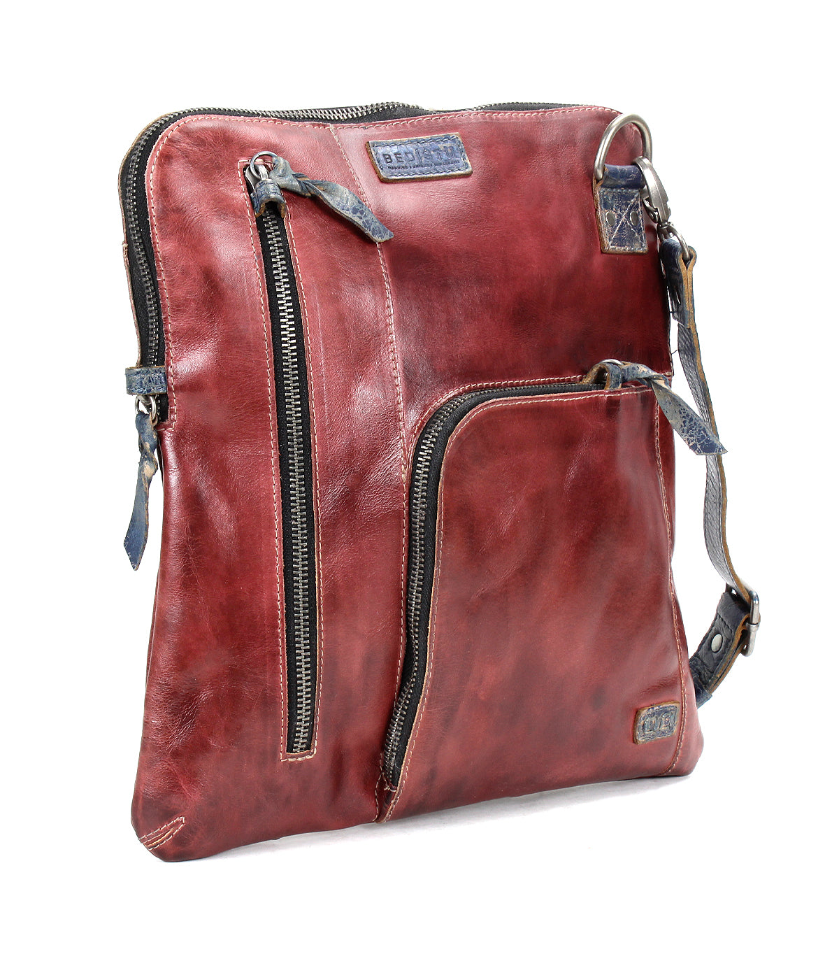 The Aiken by Bed|Stü is a square-shaped crossbody bag crafted from reddish-brown full-grain leather, featuring two zippered front pockets, a blue adjustable strap, and visible stitching.