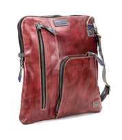 The Aiken by Bed|Stü is a square-shaped crossbody bag crafted from reddish-brown full-grain leather, featuring two zippered front pockets, a blue adjustable strap, and visible stitching.