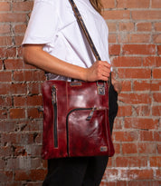 A person wearing a white t-shirt and black pants carries the Bed|Stü Aiken—a red full-grain leather crossbody bag with an adjustable strap and zippered pockets—while standing in front of a brick wall.