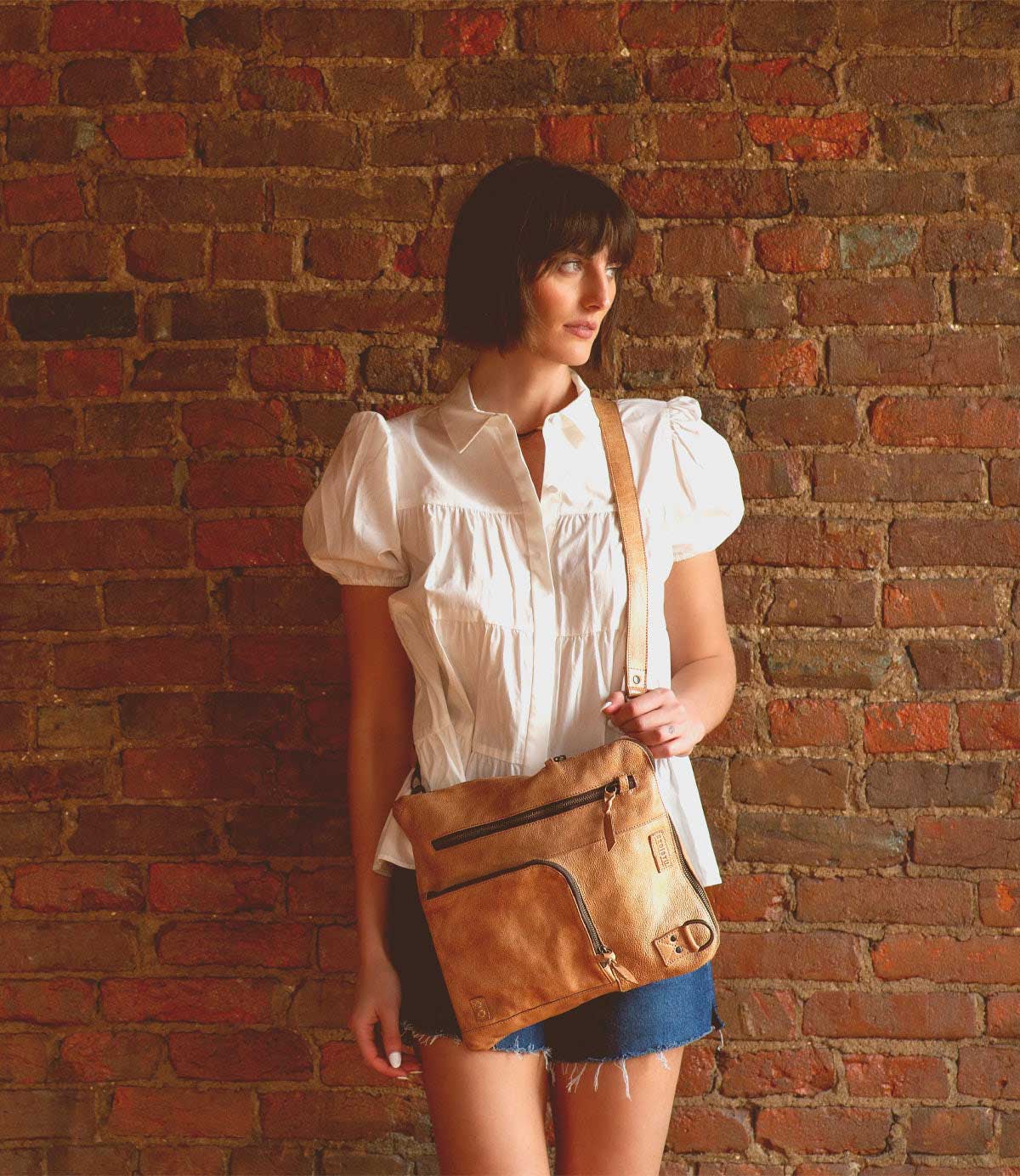 A woman in a white blouse and denim skirt stands against a brick wall, carrying the Bed|Stü Aiken crossbody bag, featuring a tan finish and an adjustable strap for comfort.