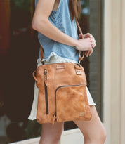 Wearing a sleeveless denim top and white skirt, the person carries the Bed|Stü Aiken—a tan full-grain leather bag featuring pockets, zippers, and an adjustable strap for comfort.