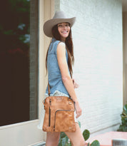 A woman in a denim vest and wide-brimmed hat stands outdoors, smiling, with the Bed|Stü Aiken tan full-grain leather bag featuring an adjustable strap slung over her shoulder.
