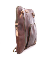 The Bed|Stü Aiken is a brown full-grain leather zippered pouch shown standing upright at an angle, featuring two zippered compartments, anti-theft security features, and a brand tag on the front.