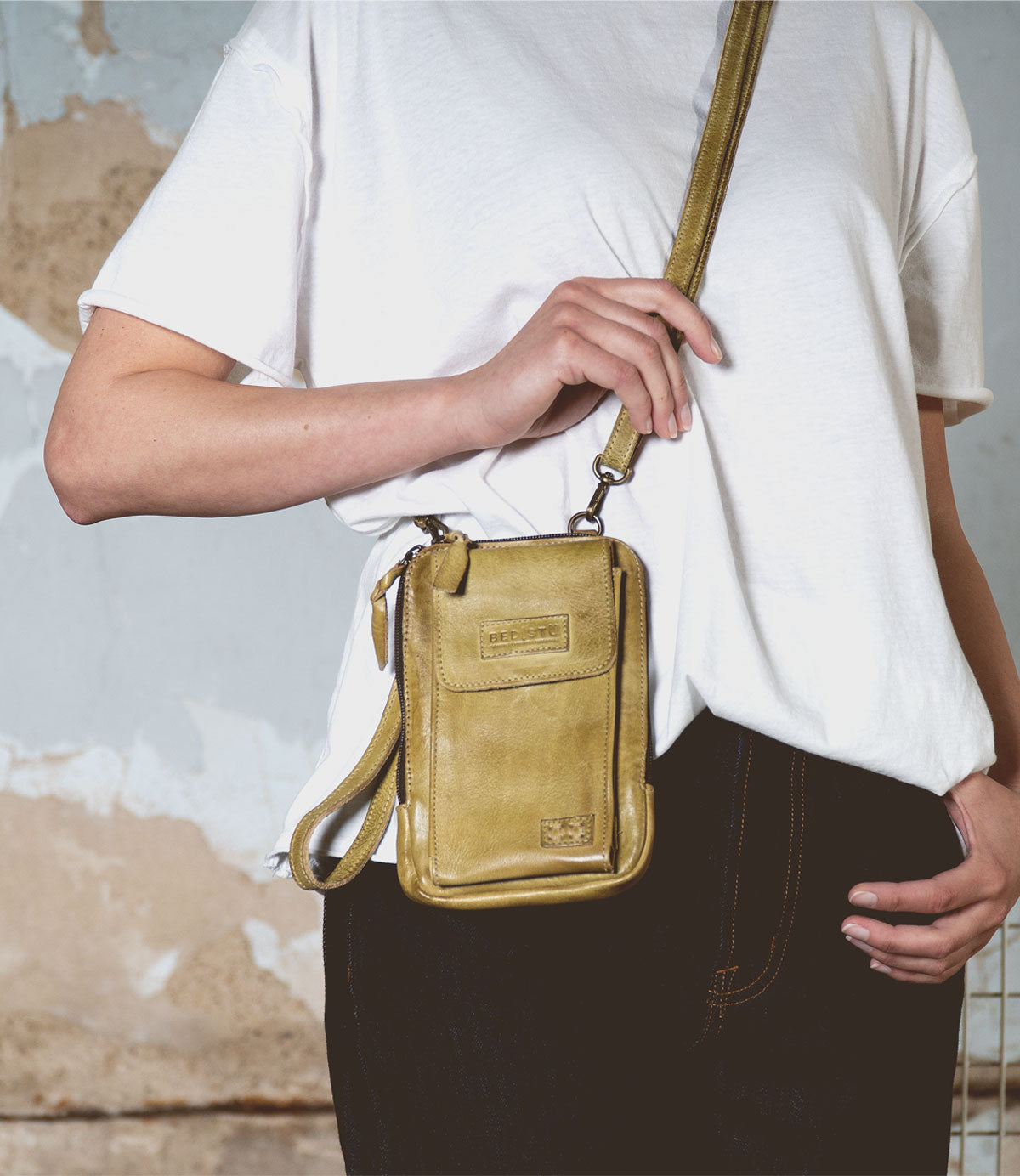 Wearing a white T-shirt and dark pants, a person holds the Bed|Stü Alelike, a compact purse-style yellow crossbody bag, against a textured wall—an ideal choice for carrying your travel essentials on the go.