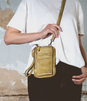 Wearing a white T-shirt and dark pants, a person holds the Bed|Stü Alelike, a compact purse-style yellow crossbody bag, against a textured wall—an ideal choice for carrying your travel essentials on the go.