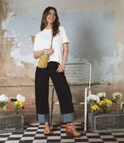 Clad in a white t-shirt and dark cuffed jeans, a woman stands smiling on a checkered floor lined with crates of white and yellow flowers, against a weathered, painted wall—her Bed|Stü Alelike purse resting effortlessly at her side, the ideal companion among her travel essentials.