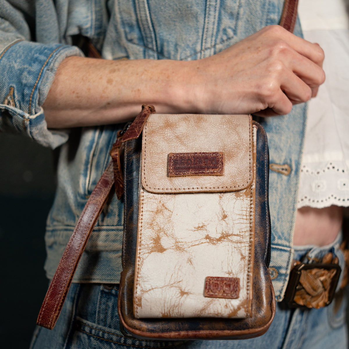 A person wearing a light denim jacket holds the Bed|Stü Alelike—a worn leather crossbody bag featuring visible marbling and a brown strap—perfect for carrying travel essentials.