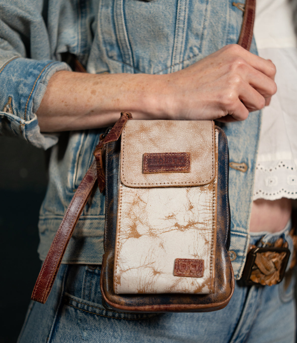 A person wearing a light denim jacket holds the Bed|Stü Alelike—a worn leather crossbody bag featuring visible marbling and a brown strap—perfect for carrying travel essentials.