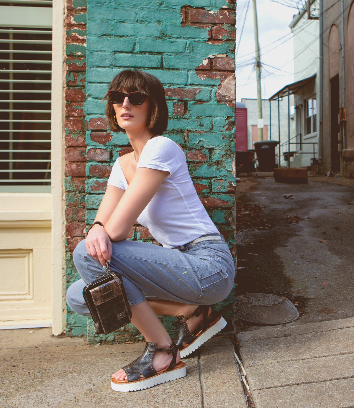 A woman with short brown hair and sunglasses squats by a brick wall, wearing a white shirt, blue jeans, and sandals, holding the Bed|Stü Alelike compact purse. An alleyway is visible in the background—her Alelike leather crossbody is perfect for travel essentials.