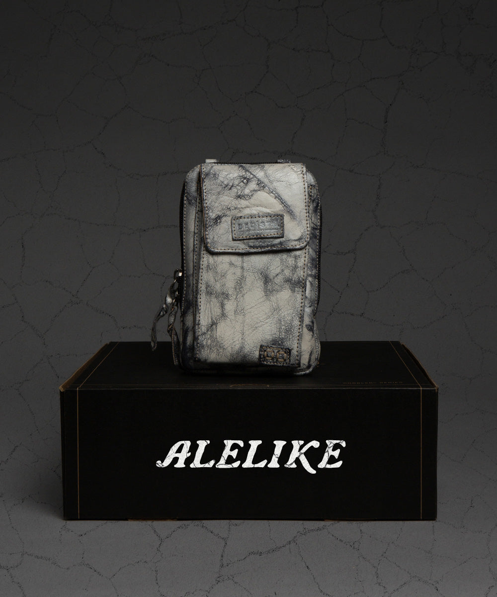 A compact purse from Bed|Stü's Alelike collection, featuring a marble pattern and zipper closure with a label, is displayed atop a black “ALELIKE” box against a cracked gray backdrop.