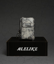 A compact purse from Bed|Stü's Alelike collection, featuring a marble pattern and zipper closure with a label, is displayed atop a black “ALELIKE” box against a cracked gray backdrop.