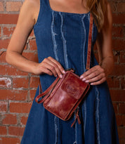 Wearing a blue denim dress, the woman holds the Bed|Stü Alelike compact leather crossbody purse against a brick wall background—perfect for keeping your travel essentials stylishly organized.