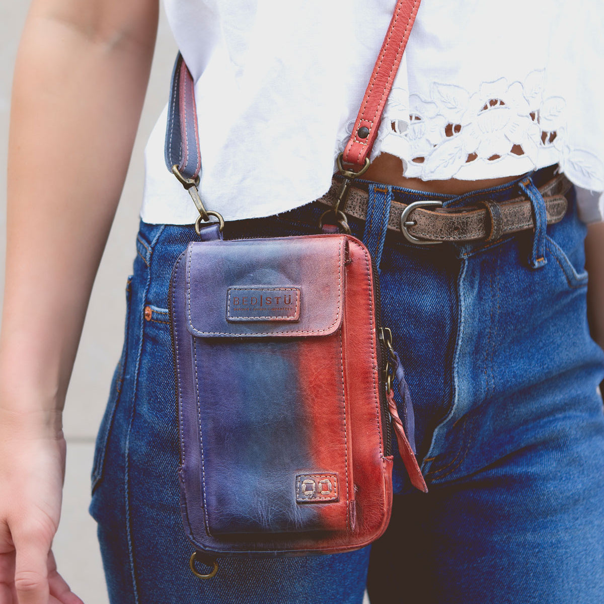 Wearing blue jeans, a white lace-detailed top, and the Alelike crossbody bag by Bed|Stü in red and blue leather—perfect for carrying travel essentials—paired with a brown belt.