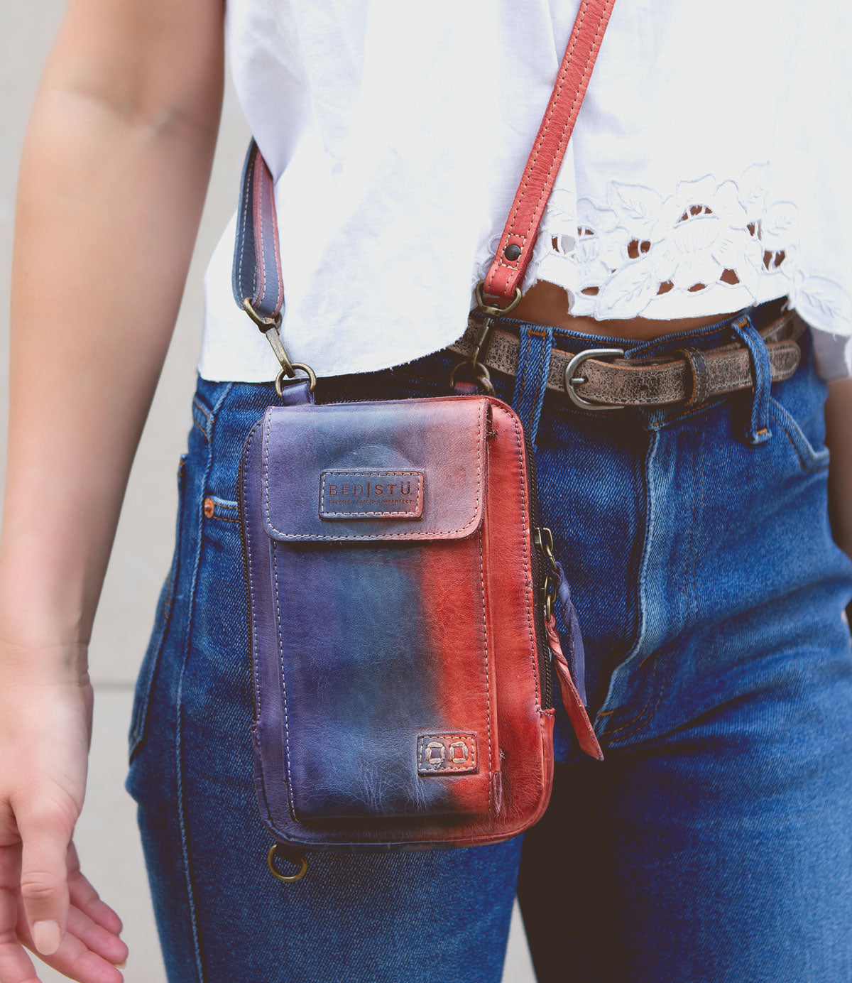 Wearing blue jeans, a white lace-detailed top, and the Alelike crossbody bag by Bed|Stü in red and blue leather—perfect for carrying travel essentials—paired with a brown belt.