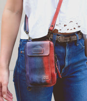 Wearing blue jeans, a white lace-detailed top, and the Alelike crossbody bag by Bed|Stü in red and blue leather—perfect for carrying travel essentials—paired with a brown belt.
