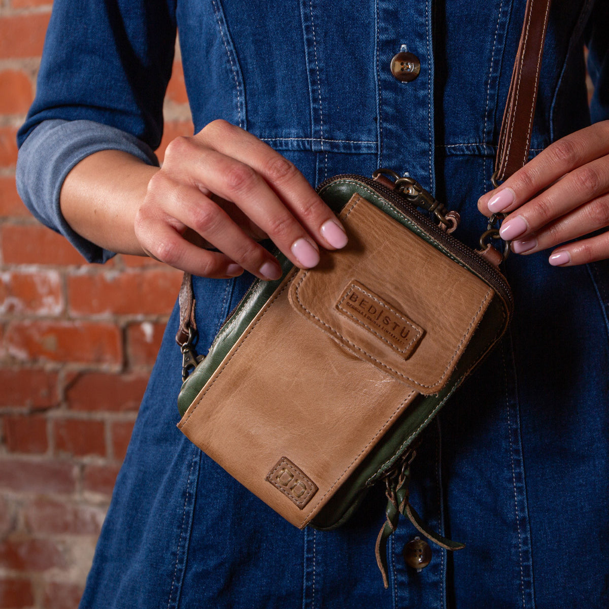 A person in a blue denim dress holds the Bed|Stü Alelike—a compact tan and green leather crossbody purse—with both hands in front of a brick wall, perfect for keeping travel essentials close at hand.