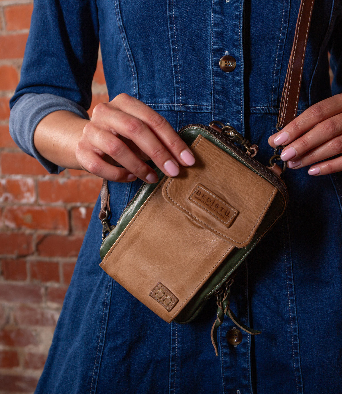 A person in a blue denim dress holds the Bed|Stü Alelike—a compact tan and green leather crossbody purse—with both hands in front of a brick wall, perfect for keeping travel essentials close at hand.