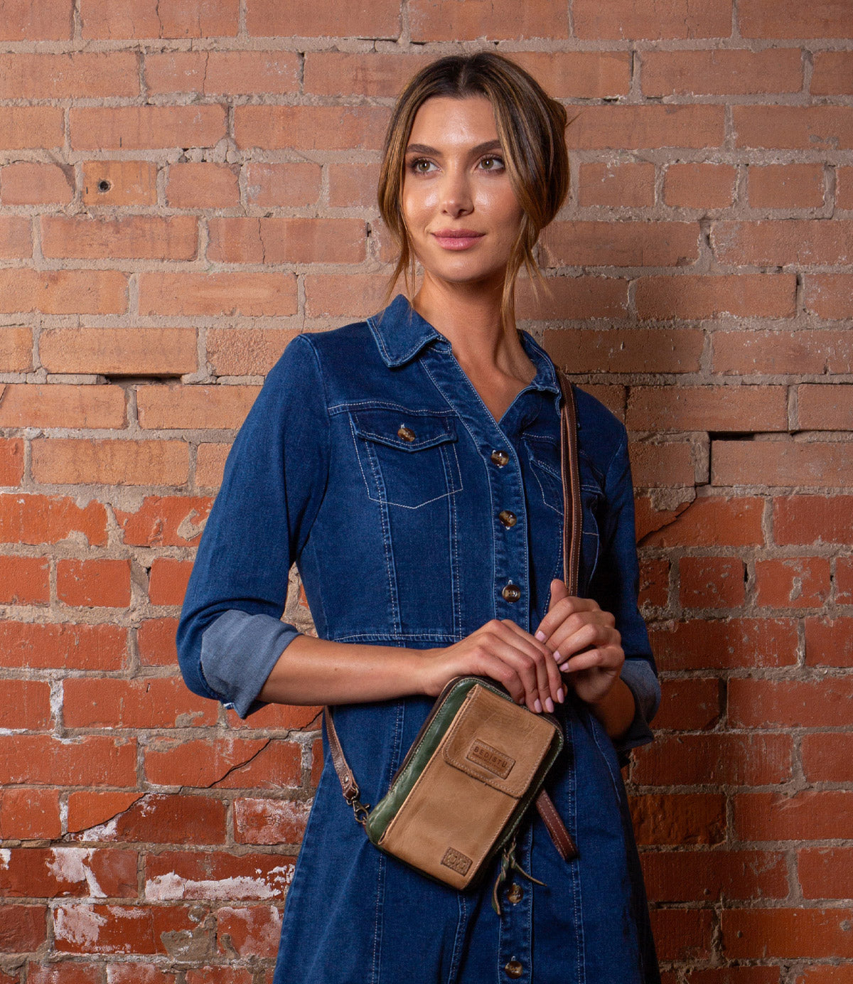 A woman in a blue denim dress stands against a brick wall, holding the Alelike by Bed|Stü—a small green and tan leather crossbody purse that's perfect for travel essentials.