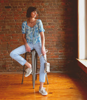 A woman in a blue floral top and light jeans sits on a metal stool by a brick wall, holding the Bed|Stü Alelike compact purse, as sunlight streams through a window onto the wooden floor.