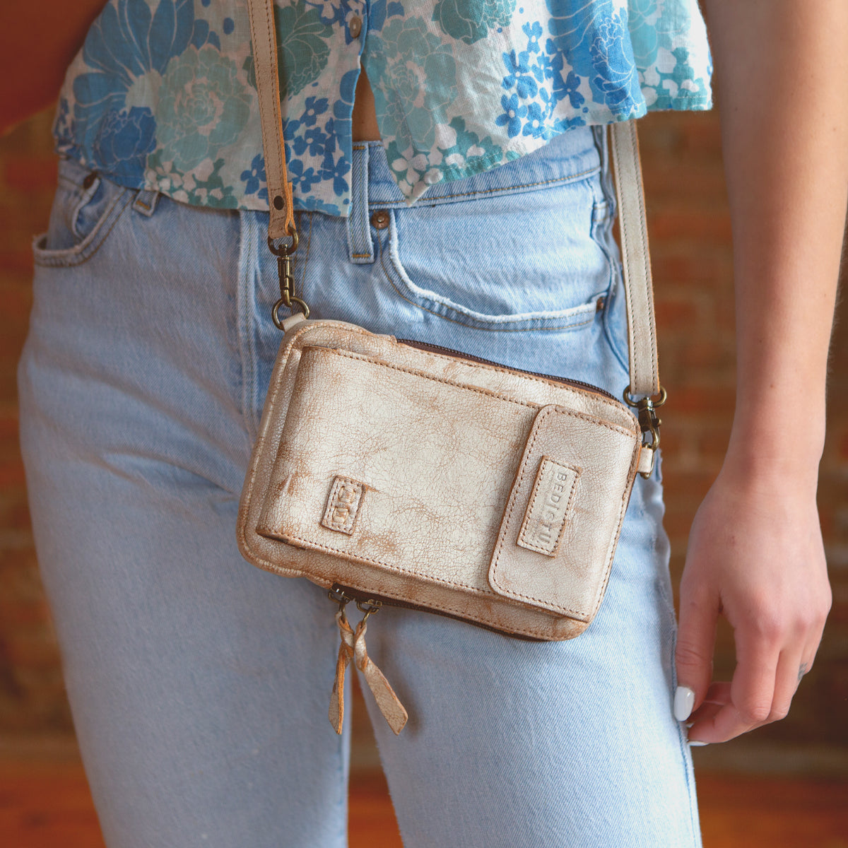 A person wearing light blue jeans, a blue floral crop top, and carrying the Bed|Stü Alelike—a compact leather crossbody purse with a zipper and strap, perfect for keeping travel essentials organized and within easy reach.