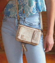 A person wearing light blue jeans, a blue floral crop top, and carrying the Bed|Stü Alelike—a compact leather crossbody purse with a zipper and strap, perfect for keeping travel essentials organized and within easy reach.