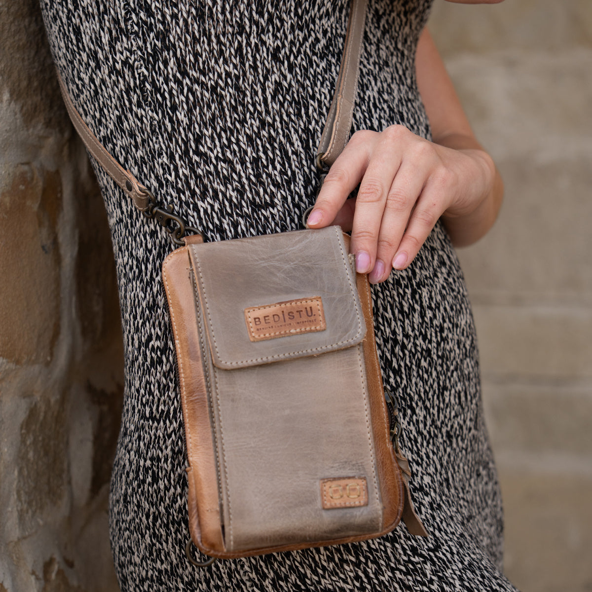 Wearing a patterned dress, a person holds the Bed|Stü Alelike—a small brown leather crossbody bag perfect for travel essentials—against their side, with a stone wall in the background.