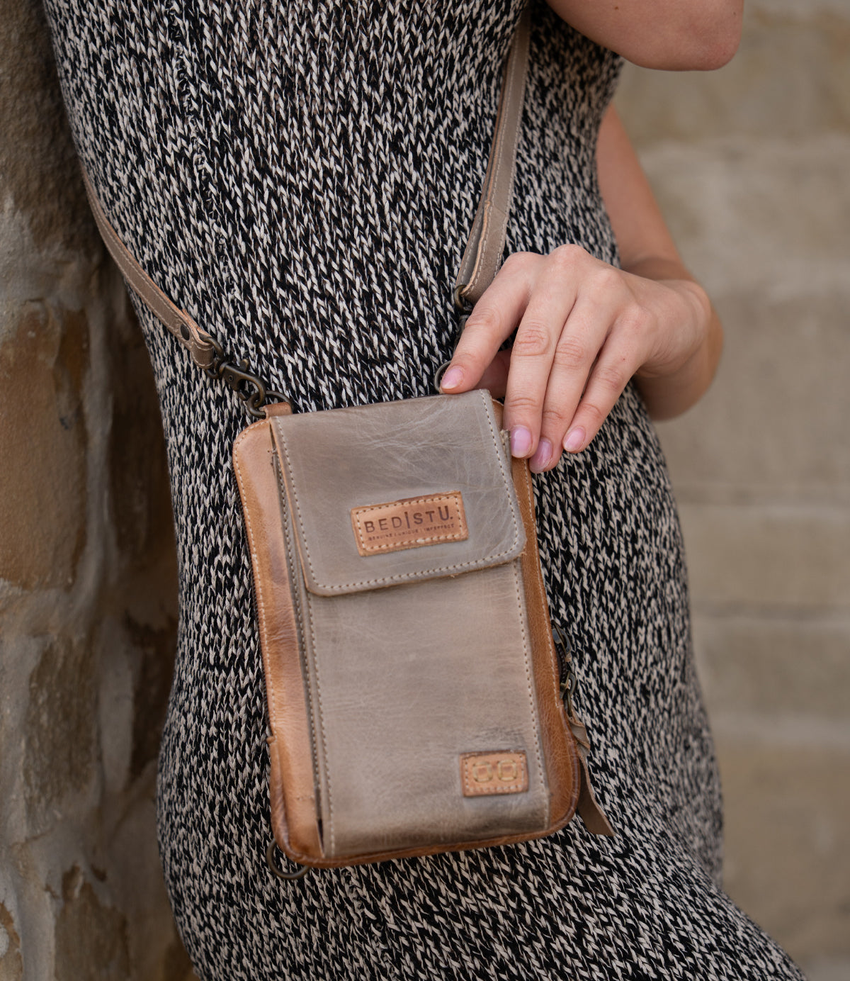Wearing a patterned dress, a person holds the Bed|Stü Alelike—a small brown leather crossbody bag perfect for travel essentials—against their side, with a stone wall in the background.