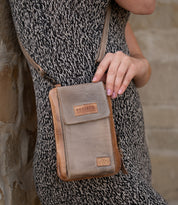 Wearing a patterned dress, a person holds the Bed|Stü Alelike—a small brown leather crossbody bag perfect for travel essentials—against their side, with a stone wall in the background.