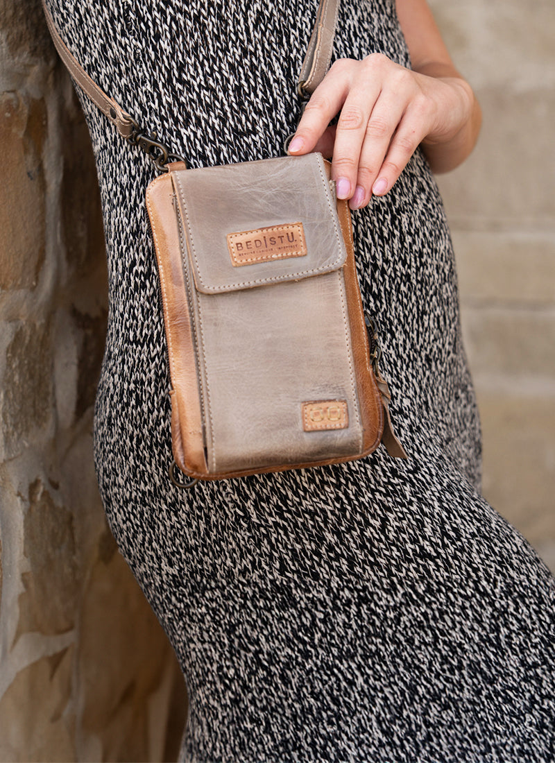 Model in a black-and-white patterned dress wears a small tan leather crossbody bag against a light stone wall.