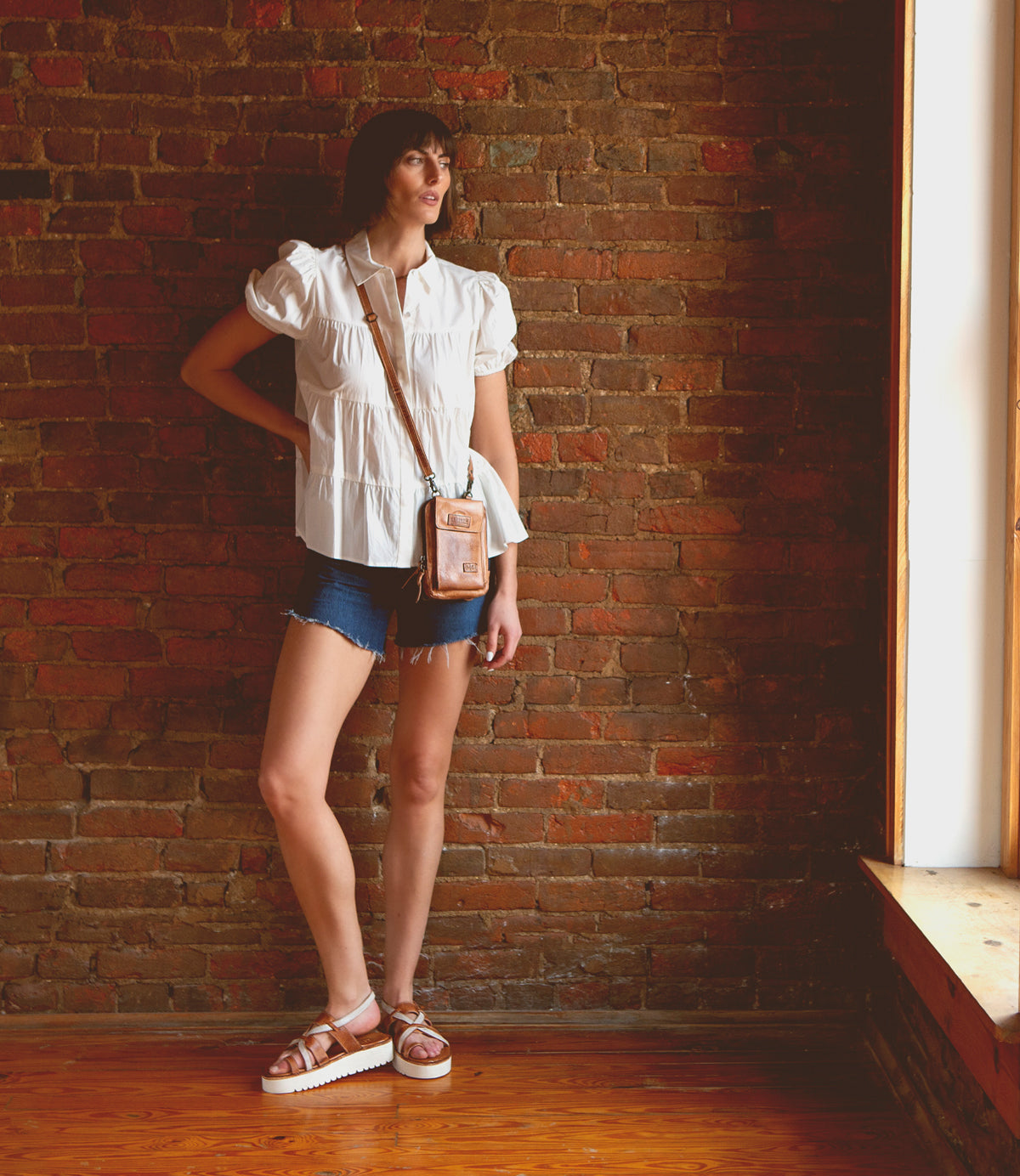 A person stands indoors by a brick wall, wearing a white blouse, denim shorts, and sandals, with the Bed|Stü Alelike leather crossbody bag filled with travel essentials slung across their body as they look toward a sunlit window.