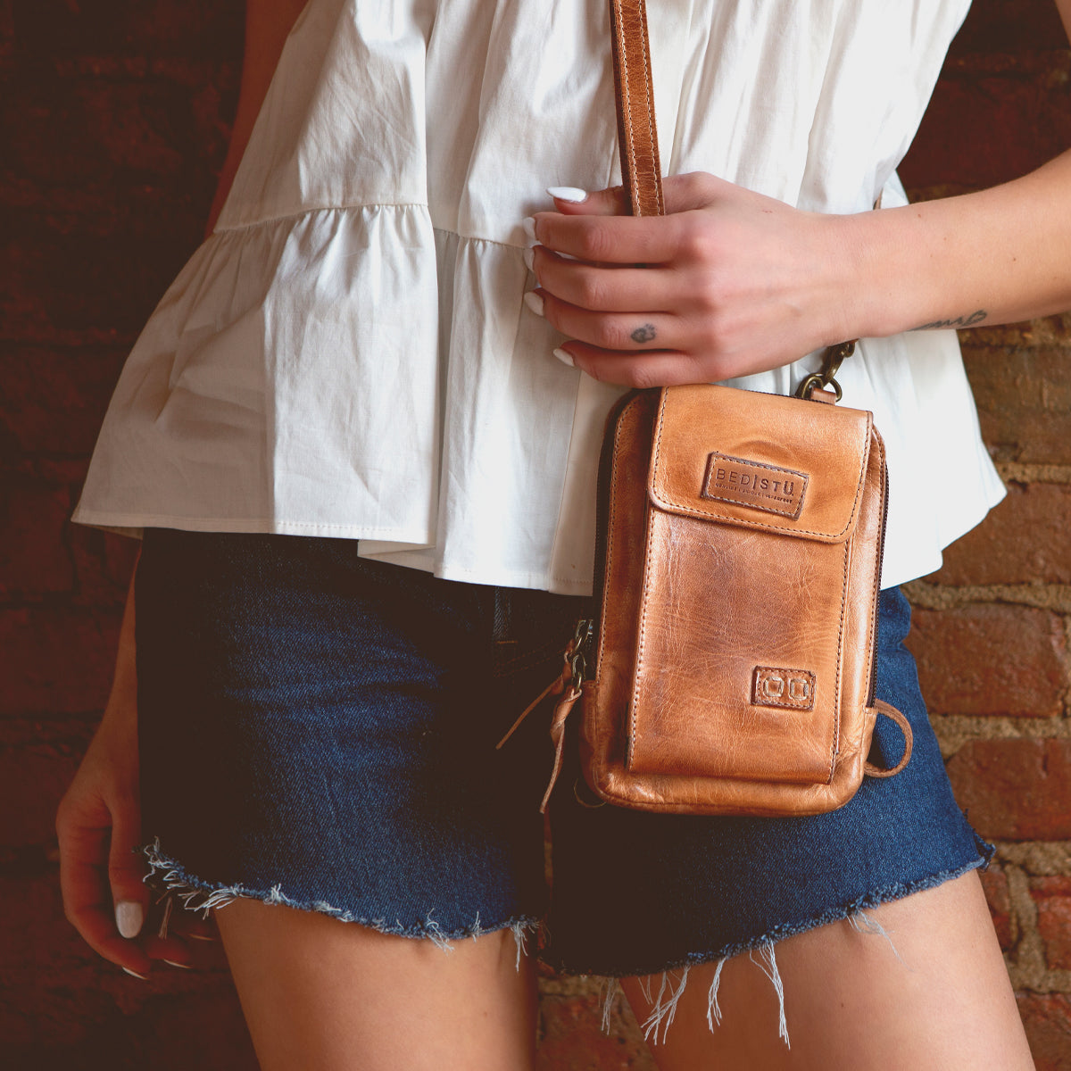 Wearing a white sleeveless top and denim shorts, the person holds the Alelike compact leather crossbody bag by Bed|Stü—perfect for carrying travel essentials—while standing against a brick wall.