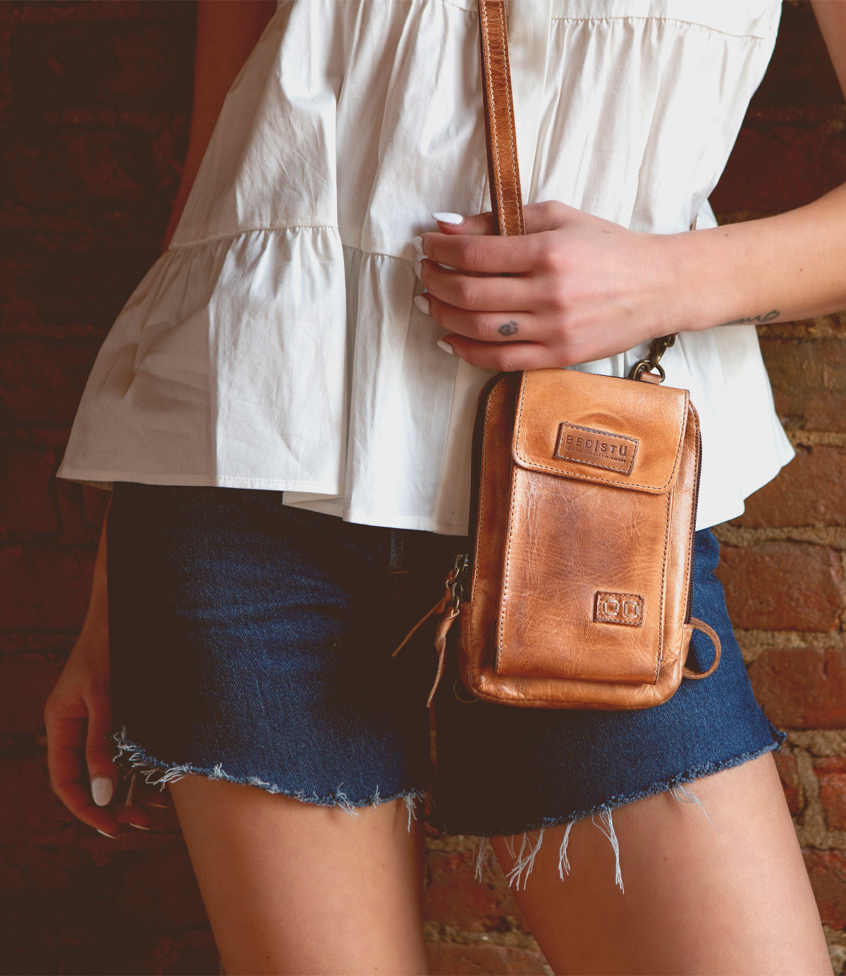 Wearing a white sleeveless top and denim shorts, the person holds the Alelike compact leather crossbody bag by Bed|Stü—perfect for carrying travel essentials—while standing against a brick wall.