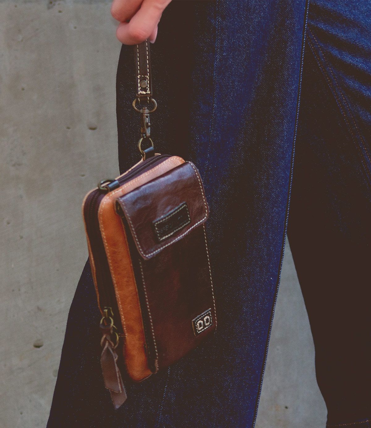 A person holding the Bed|Stü Alelike wristlet—a brown leather wallet featuring a zipper and front pocket, perfect for carrying your travel essentials—stands against a concrete wall in dark blue jeans.