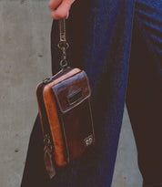 A person holding the Bed|Stü Alelike wristlet—a brown leather wallet featuring a zipper and front pocket, perfect for carrying your travel essentials—stands against a concrete wall in dark blue jeans.