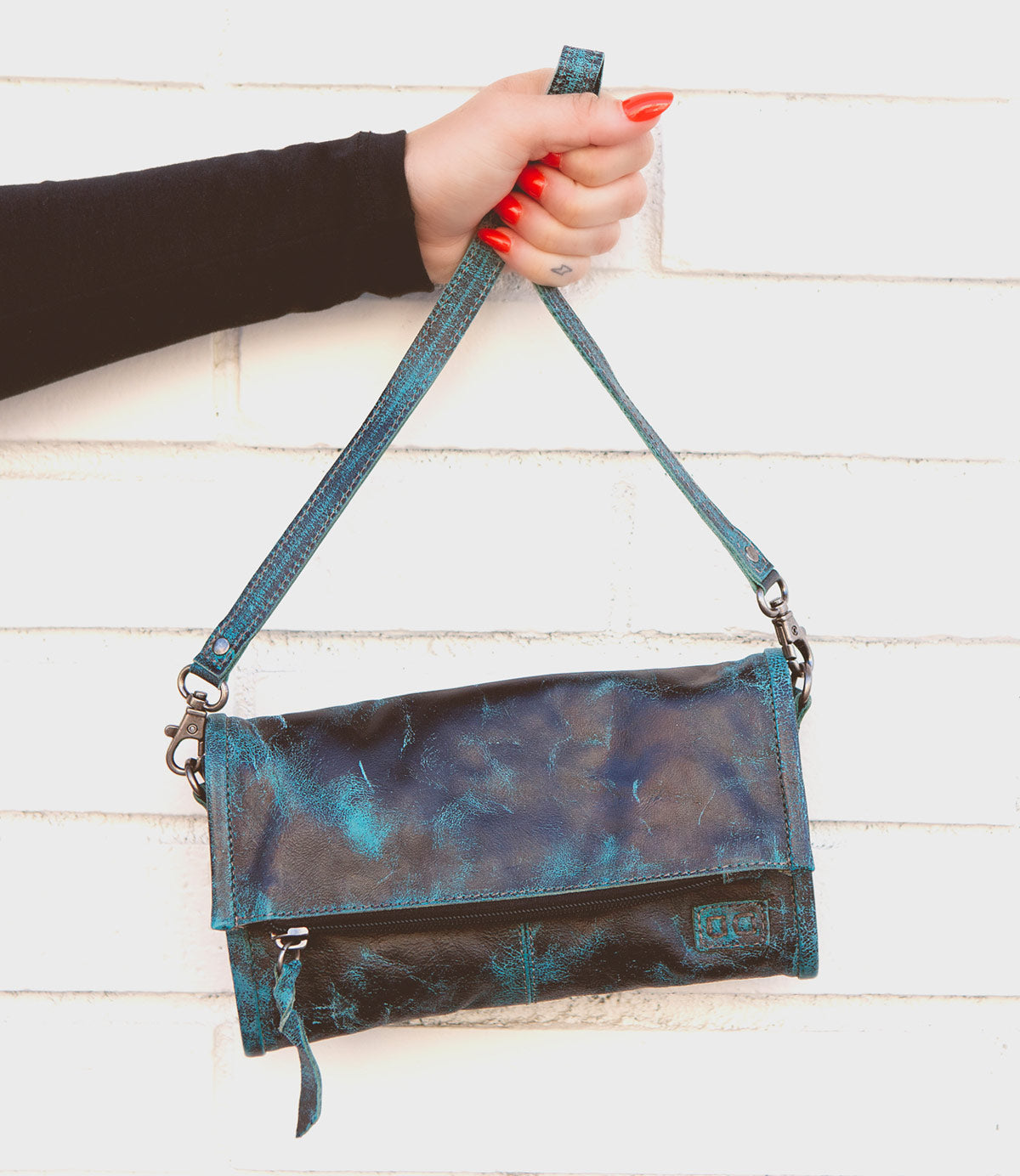 A hand with red nails holds the Amina small blue and black leather crossbody handbag by Bed|Stü by its strap against a white brick wall.
