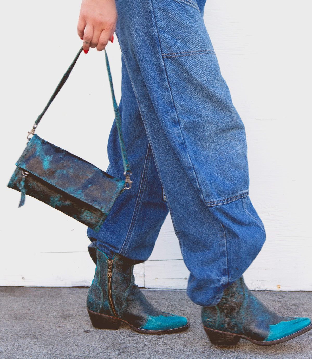 Wearing the Amina blue denim pants by Bed|Stü and teal-patterned ankle boots, this person carries a matching small crossbody purse while walking on a gray surface against a white wall.