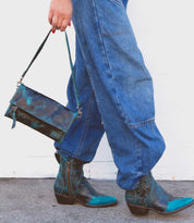 Wearing the Amina blue denim pants by Bed|Stü and teal-patterned ankle boots, this person carries a matching small crossbody purse while walking on a gray surface against a white wall.