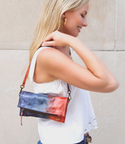 A woman with long blonde hair stands against a light beige wall, wearing a white sleeveless top and carrying the Bed|Stü "Amina" tie-dye red and blue crossbody bag.