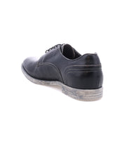 The Bed|Stü Anchiano is a black leather oxford featuring a flexible sole and visible heel wear, photographed from a rear side angle against a white background. The distressed leather finish enhances its character and showcases its well-loved appeal.