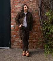 A man with long brown hair and a beard stands against a brick wall, wearing a distressed leather brown jacket, brown pants, white t-shirt, and the Anchiano tan oxford shoes by Bed|Stü, featuring a flexible sole.