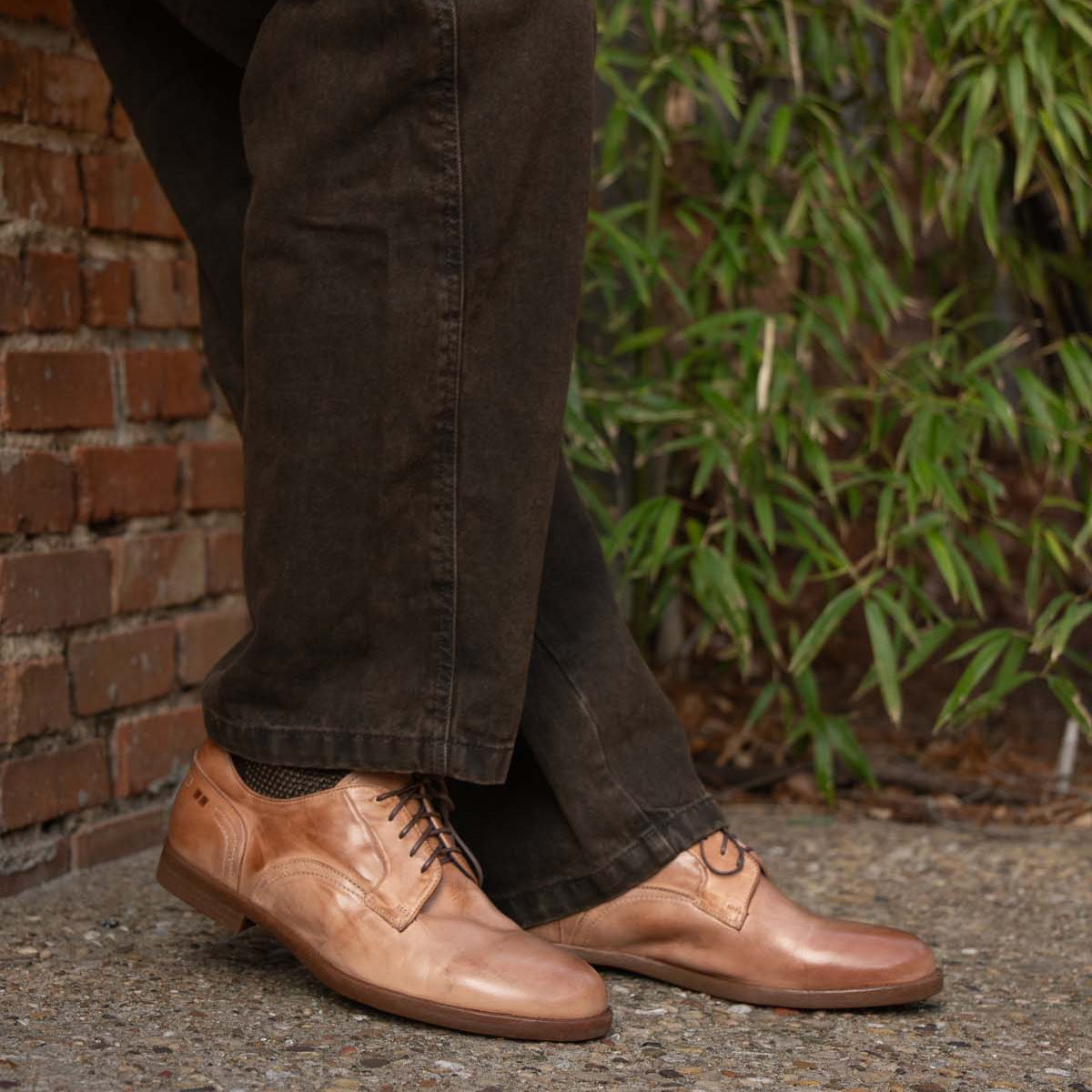 A person wearing Bed|Stü Anchiano brown oxford shoes with a flexible sole and dark pants stands on a gravel surface near a brick wall and green foliage.