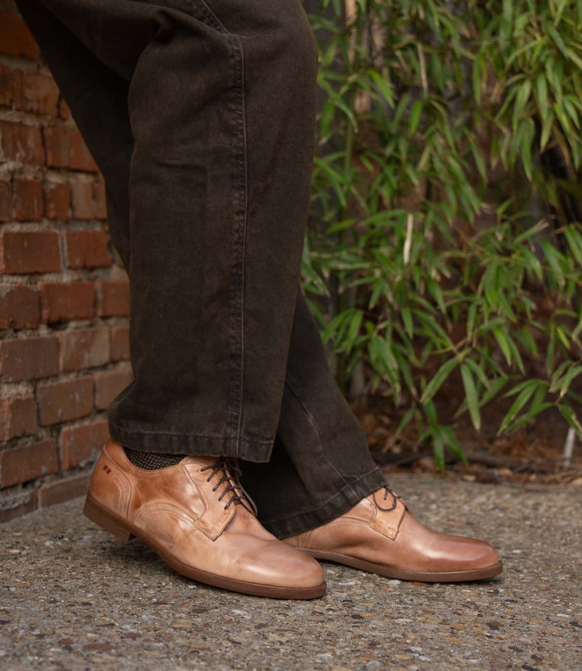 A person wearing Bed|Stü Anchiano brown oxford shoes with a flexible sole and dark pants stands on a gravel surface near a brick wall and green foliage.