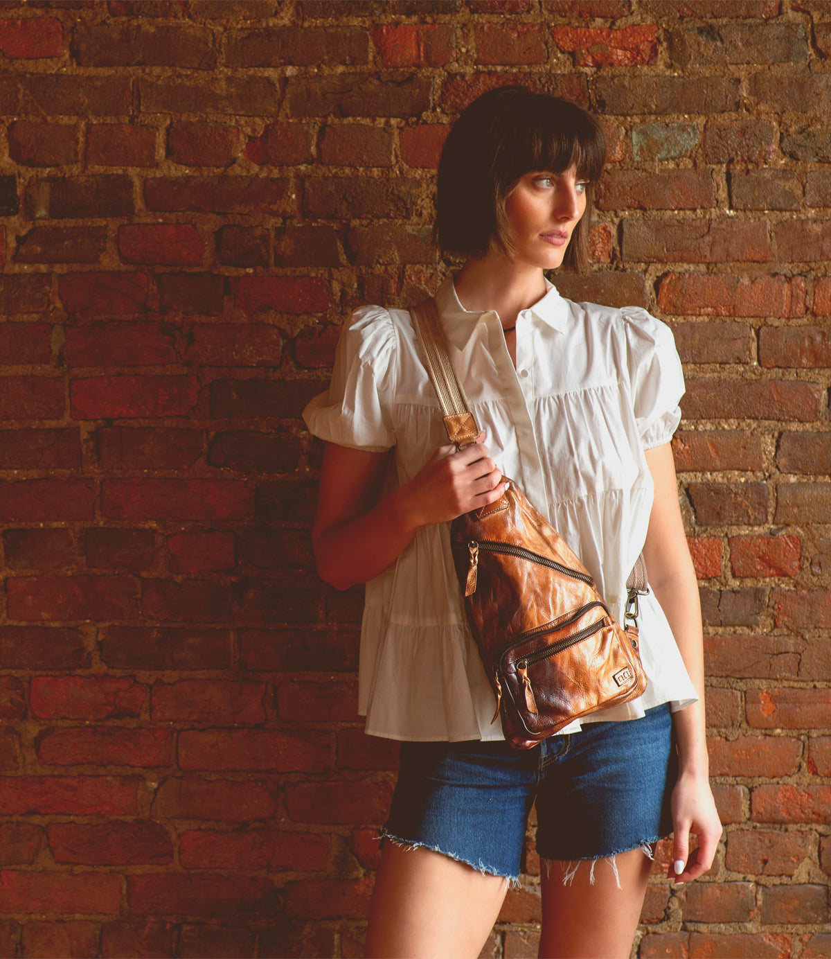 A woman in a white short-sleeve blouse and denim shorts stands against a brick wall, holding the Bed|Stü Andie—a small brown leather sling bag with an adjustable strap.