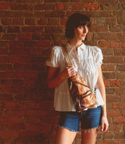 A woman in a white short-sleeve blouse and denim shorts stands against a brick wall, holding the Bed|Stü Andie—a small brown leather sling bag with an adjustable strap.
