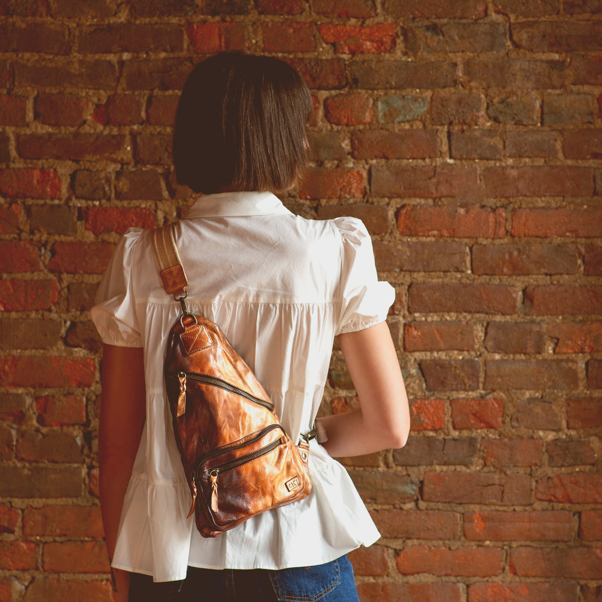 A person wearing a white blouse and denim shorts, facing a brick wall, has the Bed|Stü Andie brown leather sling bag—with an adjustable strap—on their back.