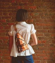 A person wearing a white blouse and denim shorts, facing a brick wall, has the Bed|Stü Andie brown leather sling bag—with an adjustable strap—on their back.